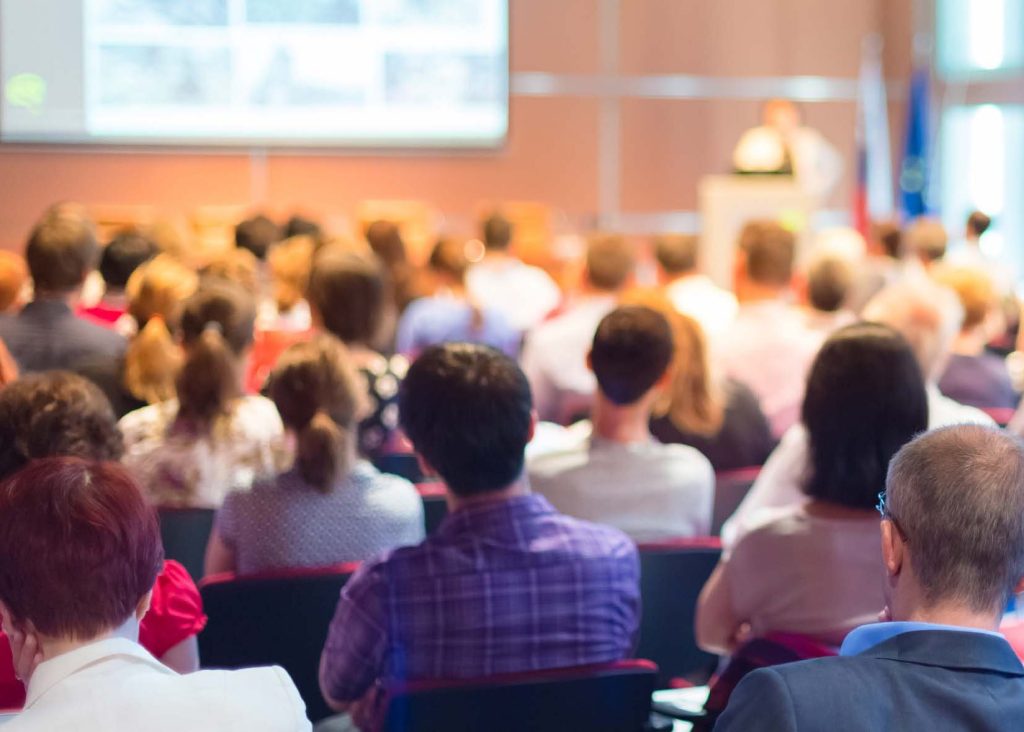 A photograph of a room full of people who are seated and watching a person making a presentation.  
